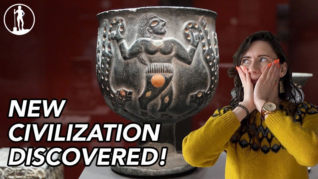 Jiroft: Iran's Lost Civilization - an interestingly sculpted pot with a figure of a dancing person with an orange stone on their belly, and a surprised person standing to its screen right.